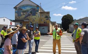 Dan Burden stands at pedestrian crossing with several attendees, in front of Blue Lake downtown mural.
